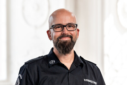 Prof. Dr. Heiko Paulheim trägt eine dunkle Brille und ein dunkles Hemd, er hat eine Glatze, einen dunklen Bart und Ohrringe. 