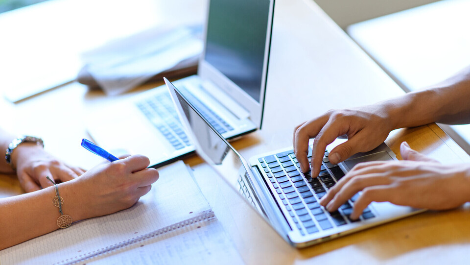 Arbeitsplatz Zwei Laptops stehen versetzt auf einem Tisch. Am rechten Laptop sind zwei Hände an der Tastatur. Gegenüber davon schreibt eine Person auf einen Block.