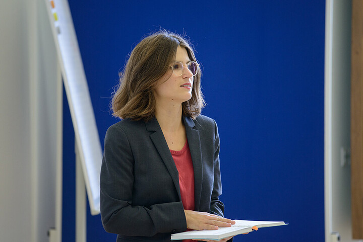 A woman is holding an open book in her hands, behind her is a flipchart. She is wearing glasses and a dark blazer with a red blouse.