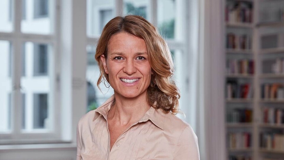 Portrait von Nadine Klass. Sie trägt eine beige farbene Bluse und steht lächelnd vor einem Fenster.