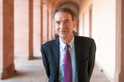 Portrait von Stefan Reichelstein. Er trägt ein weißes Hemd, eine lila Krawatte und einen schwarzen Blazer und steht in einem Torbogen.