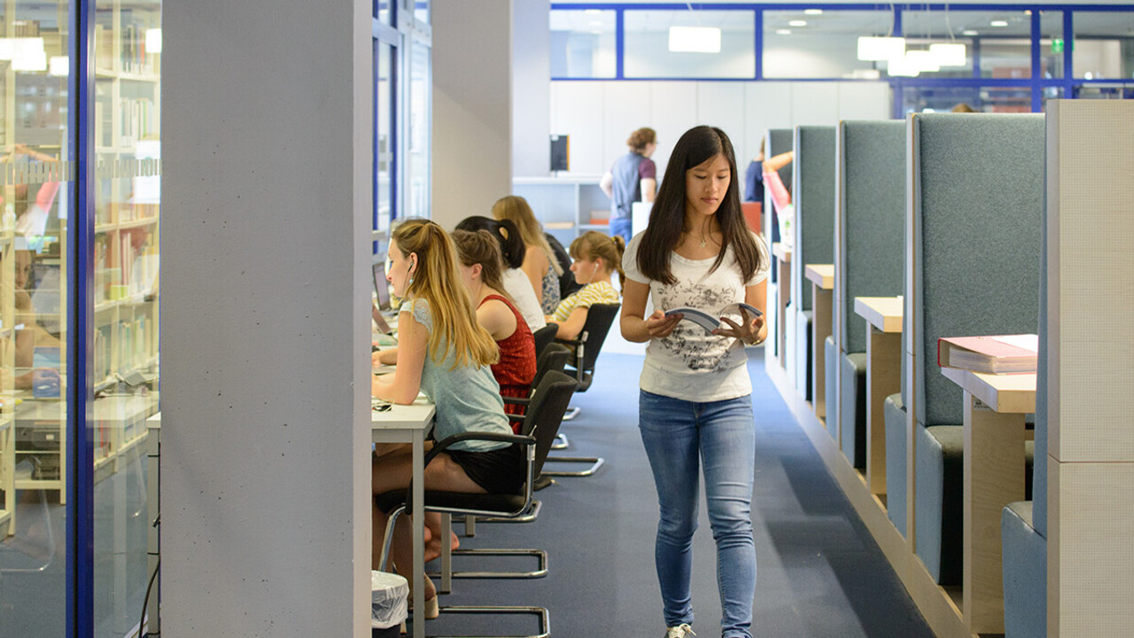 Eine Studierende läuft durch den Gruppenbereich der A5-Bibliothek und liest dabei in einem aufgeschlagenen Buch.