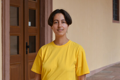 Porträt von der studierenden Person Fine Giebler (they/them). They trägt ein gelbes T-Shirt und hat kurze, dunkelbraune Haare. Im Hintergrund ist eine braune Eingangstür des Schlosses zu sehen. Link: Instagram-Post myUniMA story Juli 2022