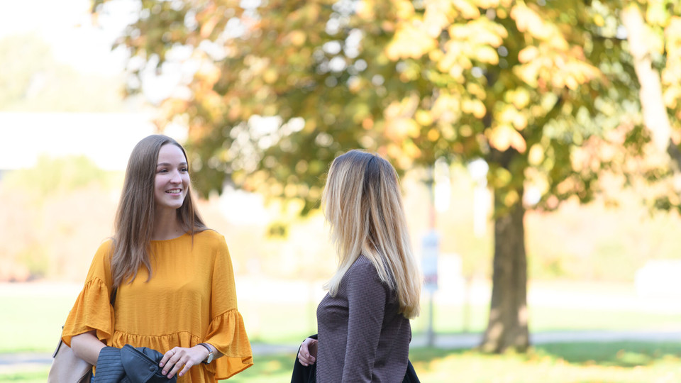 Zwei Studierende lächeln. Im Hintergrund ist ein großer Baum. 