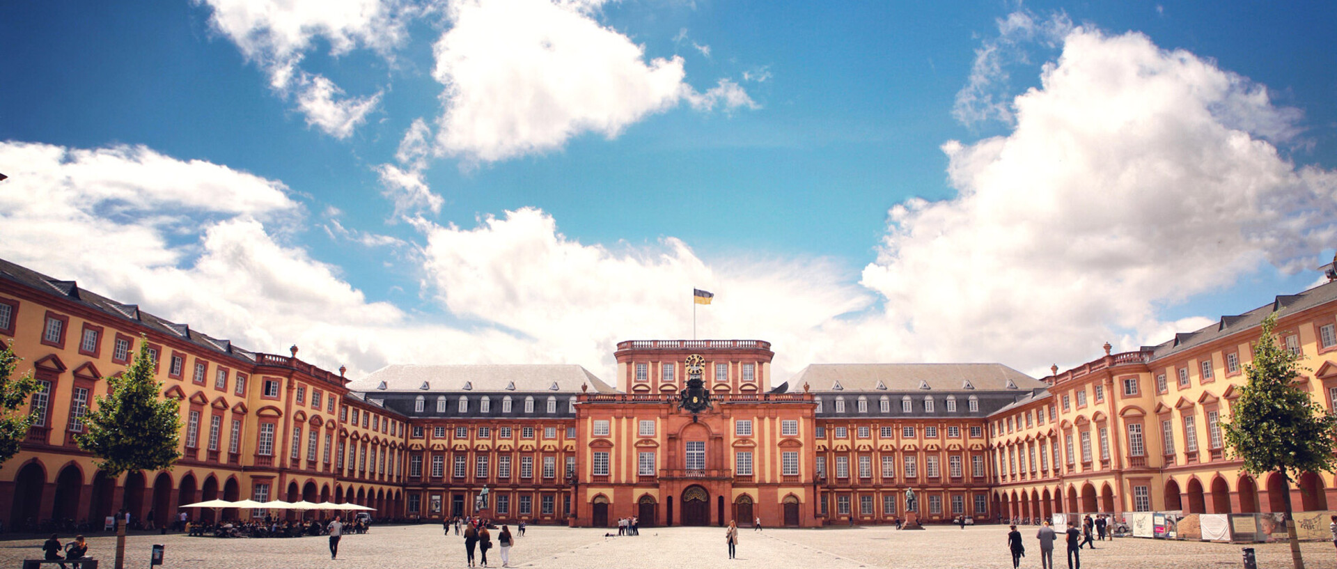 Das Barock-Schloss der Universität Mannheim steht unter blauem Himmel. Es ist von unzähligen Fenstern, rotem Sandstein und einer gelben Fassade geprägt.