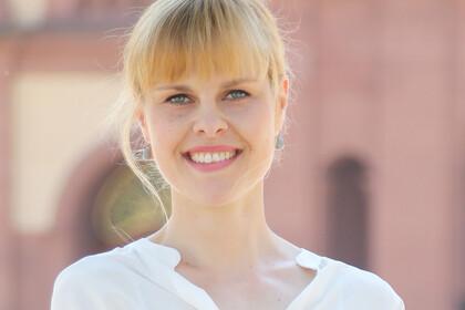 Portrait von Lea Tochtermann. Sie trägt eine weiße Bluse und steht lächelnd vor dem Schloss.
