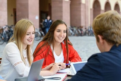 Drei Studierende sitzen auf dem Ehrenhof. Die Person links hat lange blonde Haare und trägt ein weißes Oberteil. Die Person in der Mitte hat lange braune Haare und trägt ein weißes Oberteil mit einer roten Jacke. Die Person rechts hat kurze helle Haare und trägt ein blaues Oberteil. Sie sitzen vor ihren Laptops und Unterlagen.
