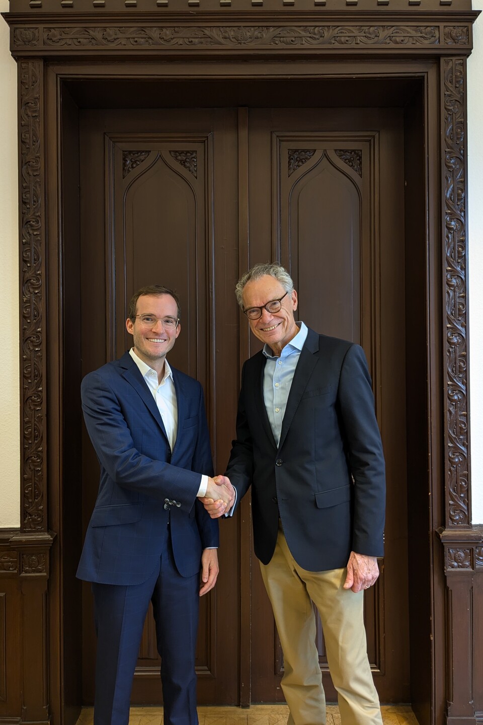 Dekan Lutz und Prof. Reinwald stehen vor einer dunklen Holztür und schütteln sich die Hände. Beide Lächeln in die Kamera.