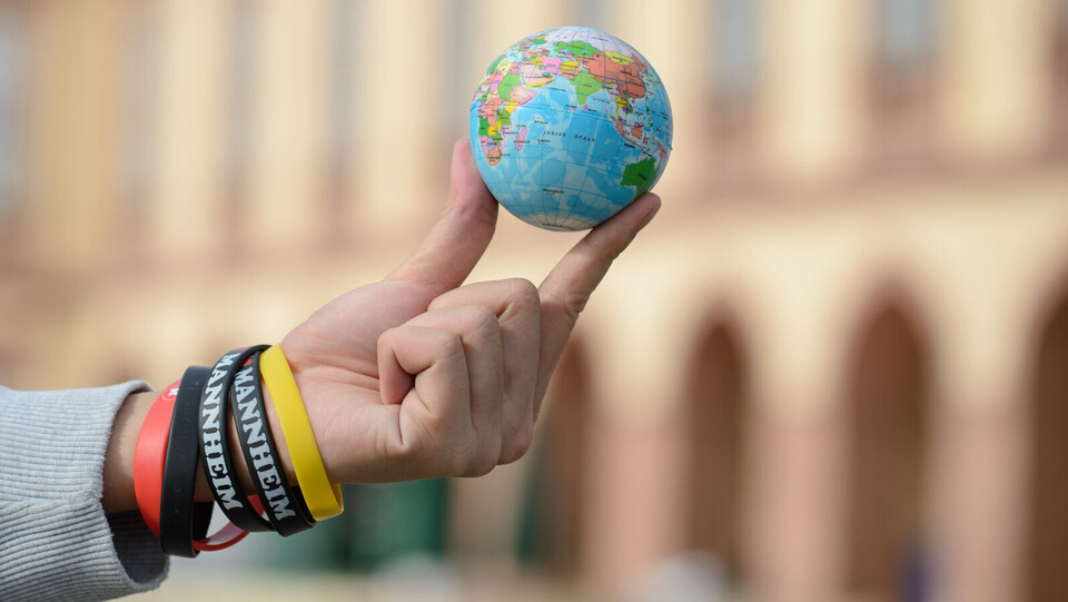 Eine Hand hält einen kleinen Weltkugelball mit zwei Fingern in die Luft.