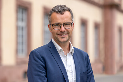 Prof. Debus steht vor dem Schloss und lächelt milde. Er trägt einen blauen Anzug und ein weißes Hemd. 