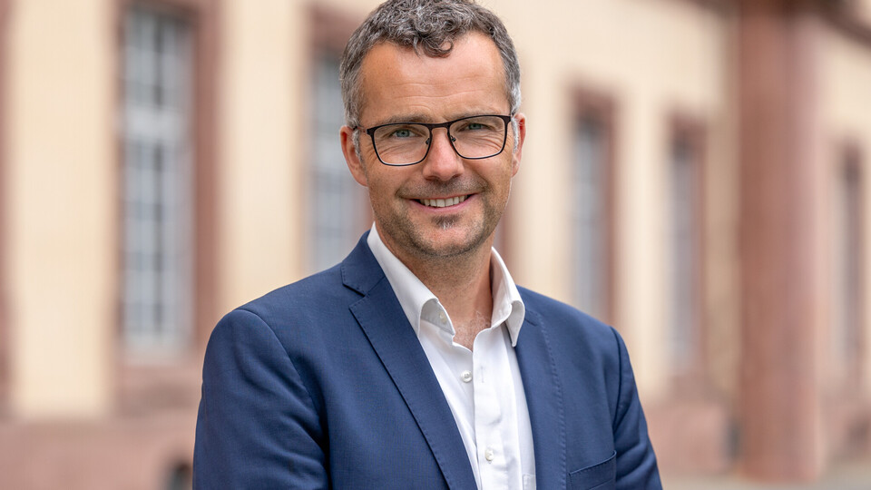 Prof. Debus steht vor dem Schloss und lächelt milde. Er trägt einen blauen Anzug und ein weißes Hemd. 