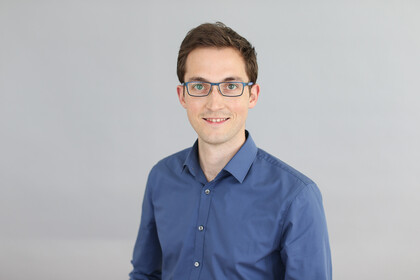 Portrait von Andreas Gerster. Er trägt ein blaues Hemd sowie eine Brille und lächelt.