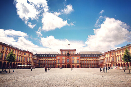 Blick auf die Universität Mannheim und den gesamten Ehrenhof. Der Himmel ist blau und bewölkt. Ein paar Menschen laufen herum.