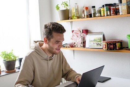 Ein Studierender hat kurze braune Haare und trägt einen beigen Kapuzenpullover. Er sitzt vor einem Laptop und macht sich Notizen.