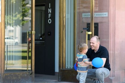 Ein junger Mann kniet vor einem kleinen Jungen und lacht diesen an. Im Hintergrund ist der Haupteingang und die Pforte der Universität zu erkennen. Link: Instagram Post Studieren mit Kind