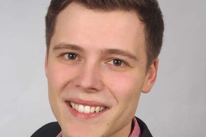 Michael Schlechtinger hat kurze dunkle Haare und trägt ein rot-kariertes Hemd unter einem dunklen Blazer. Er steht vor einer weißen Wand.