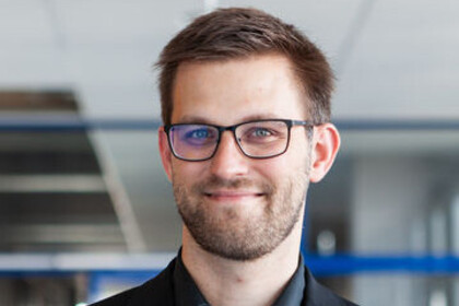 Portrait von Stefan Janke. Er trägt ein schwarzes Hemd sowie einen schwarzen Blazer und steht lächelnd in einem Uni-Gebäude.
