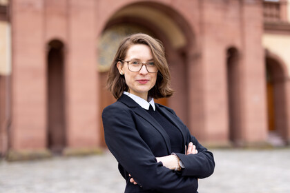 Portrait von Svenja Behrend. Sie trägt einen schwarzen Blazer und steht vor einem teil des Schlosses.
