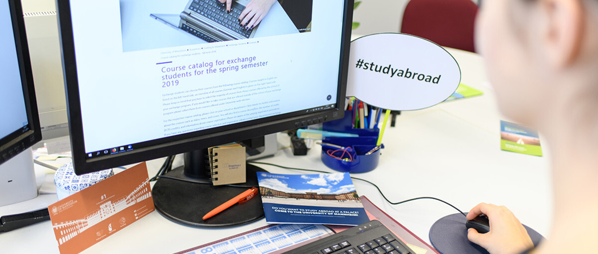 A person is sitting in front of a computer screen and looking at a website about studying abroad. On the desk are various objects such as pens, a cup and a sign saying '#studyabroad'.