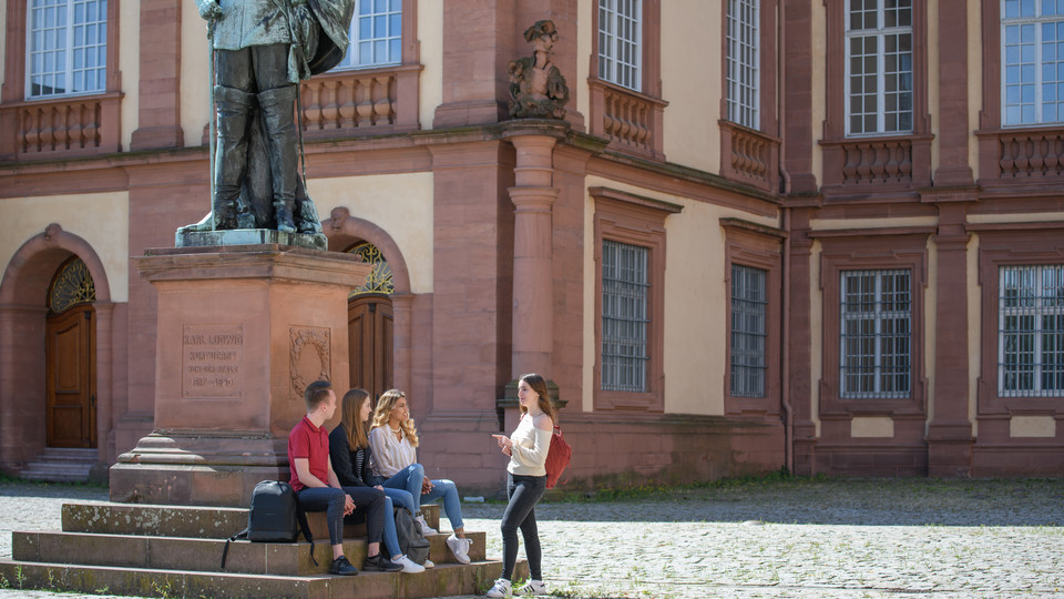 Vier Studierende sitzen im Ehrenhof vorm Schloss und einer Statue