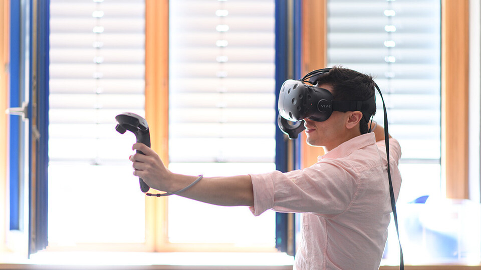 Ein Mann trägt eine Virtual Reality Brille, hält in jeder Hand einen Controller und spielt damit ein Spiel.