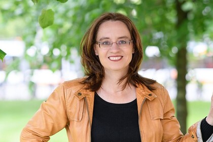 Portrait von Michele Tertilt. Sie trägt ein schwarzes Shirt und eine orange-farbige Jacke. Sie steht auf einer Grünfläche neben einem Baum.