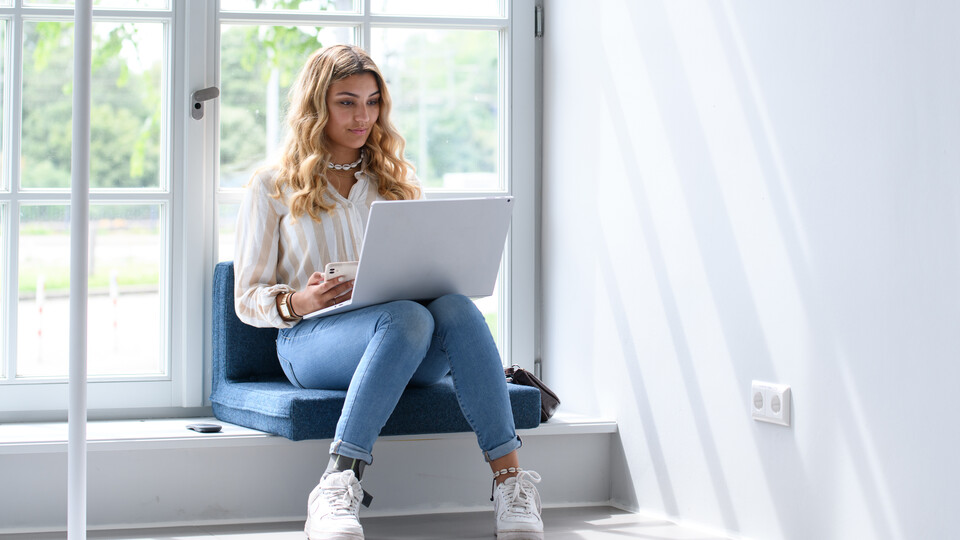 Studierende sitzt in einem große, hellen Fenster und schaut auf ihr Laptop.