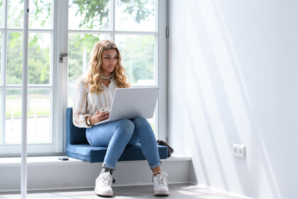 Studierende sitzt in einem große, hellen Fenster und schaut auf ihr Laptop.