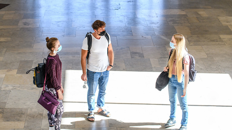 Drei Studierende stehen mit Abstand zu einander. Sie tragen Masken. 