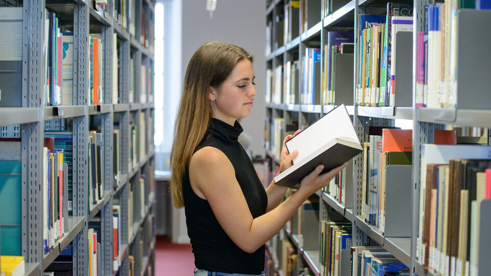 Eine Studentin steht zwischen zwei Bücherregalen und liest in einem Buch.