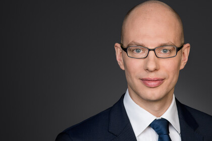 Portrait von Nils Schaks. Er trägt ein weißes Hemd, eine dunkelblaue Krawatte sowie einen dunkelblauen Blazer und steht vor einer dunkelgrauen Wand. 