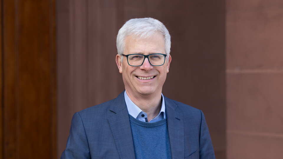 Portrait von Eckhard Janeba. Er trägt ein hellblaues Hemd, einen blauen Pullover sowie einen blauen Blazer und steht lächelnd vor einem Gebäude.