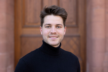 Felix Rusche. He is wearing a black sweater and is standing in front of brown door.