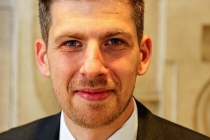 Dimitrios Linardatos hat kurze braune Haare und einen Dreitagebart. Er trägt einen schwarzen Anzug mit einer rot-blau karierten Krawatte.