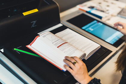 Eine Person, deren Hände im Bild sind, benutzt einen Scanner in der Bibliothek. Auf der Scanfläche liegt ein aufgeschlagenes Buch.