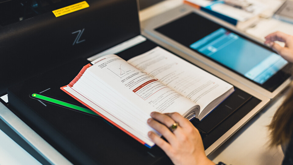 Eine Person, deren Hände im Bild sind, benutzt einen Scanner in der Bibliothek. Auf der Scanfläche liegt ein aufgeschlagenes Buch.