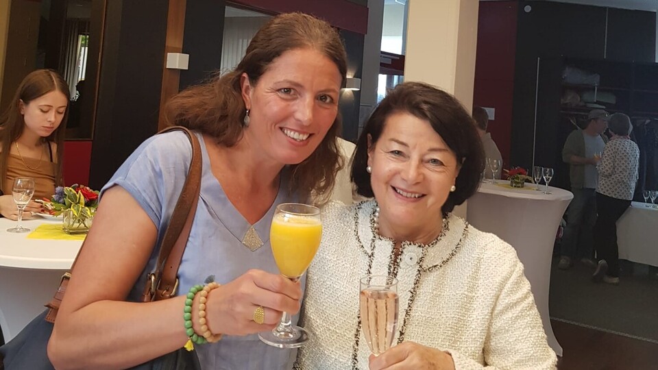 zwei Frauen lachen mit einem Glas Sekt und Organgensaft in die Kamera