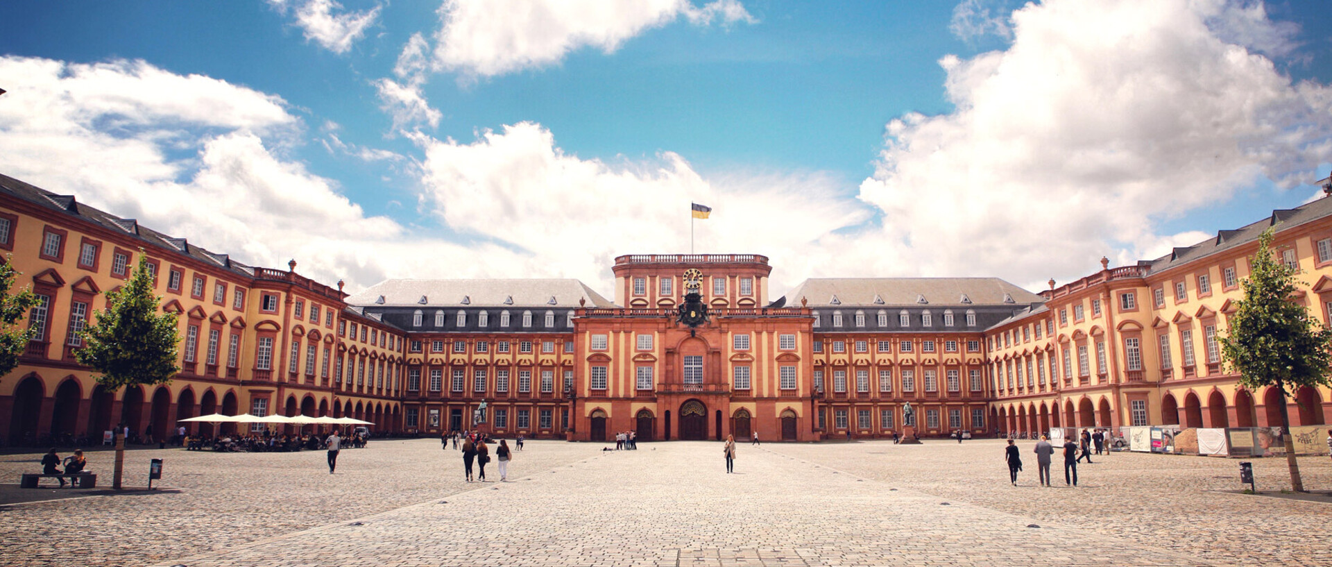 Das Barock-Schloss und der Ehrenhof der Universität Mannheim unter strahlend blauem Himmel. Das Schloss ist von unzähligen Fenstern, rotem Sandstein und einer gelben Fassade geprägt.