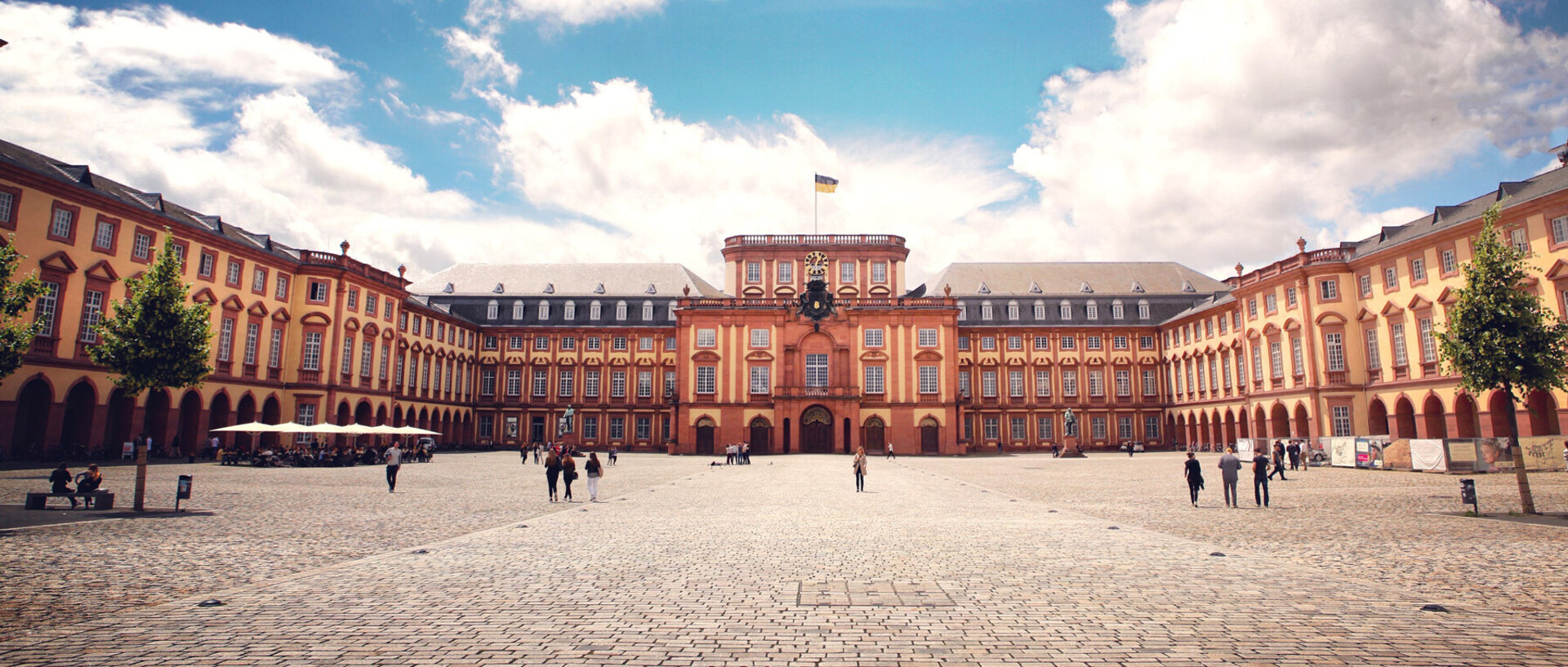 Das Barock-Schloss und der Ehrenhof der Universität Mannheim unter strahlend blauem Himmel. Das Schloss ist von unzähligen Fenstern, rotem Sandstein und einer gelben Fassade geprägt.