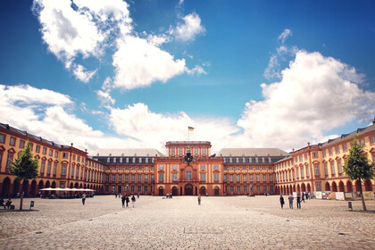 Das Barock-Schloss und der Ehrenhof der Universität Mannheim unter strahlend blauem Himmel. Das Schloss ist von unzähligen Fenstern, rotem Sandstein und einer gelben Fassade geprägt.