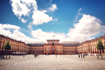 Das Barock-Schloss und der Ehrenhof der Universität Mannheim unter strahlend blauem Himmel. Das Schloss ist von unzähligen Fenstern, rotem Sandstein und einer gelben Fassade geprägt.