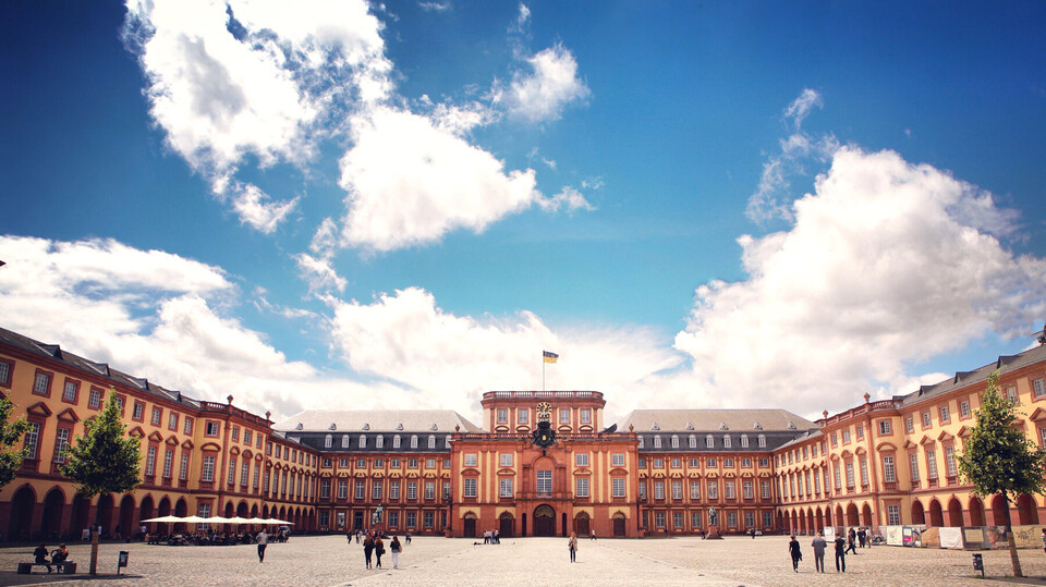 Das Barock-Schloss und der Ehrenhof der Universität Mannheim unter strahlend blauem Himmel. Das Schloss ist von unzähligen Fenstern, rotem Sandstein und einer gelben Fassade geprägt.