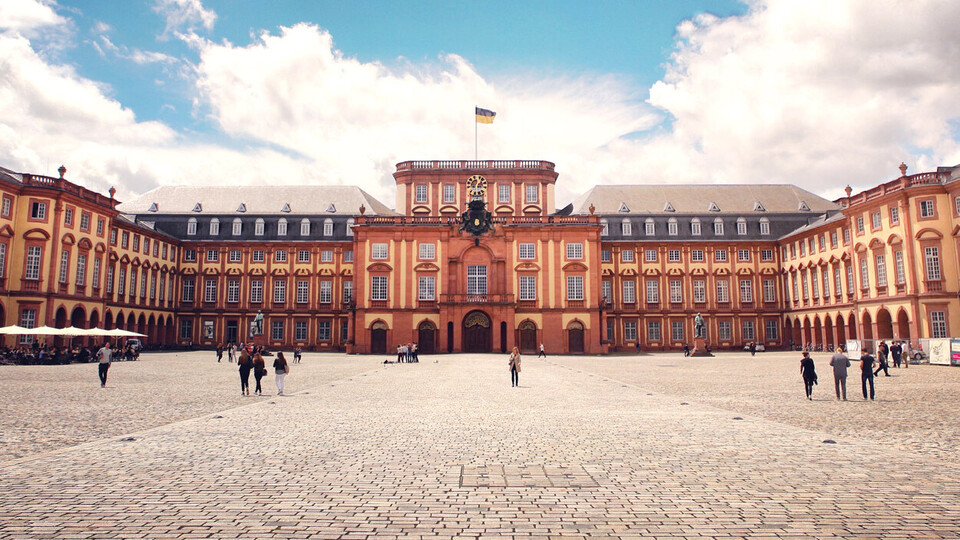 Das Barock-Schloss und der Ehrenhof der Universität Mannheim unter strahlend blauem Himmel. Das Schloss ist von unzähligen Fenstern, rotem Sandstein und einer gelben Fassade geprägt.