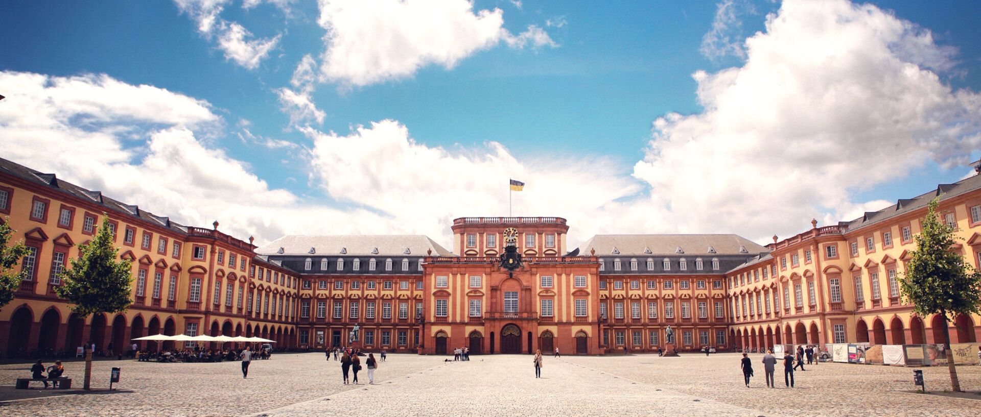 Das Barock-Schloss und der Ehrenhof der Universität Mannheim unter strahlend blauem Himmel. Das Schloss ist von unzähligen Fenstern, rotem Sandstein und einer gelben Fassade geprägt.