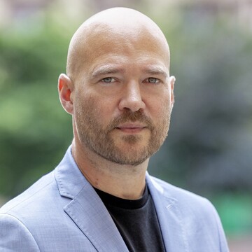 Porträtfoto von Prof. Richard Traunmüller. Er trägt ein hellblaues Sakko, ein schwarzes Shirt, er hat einen Bart. 