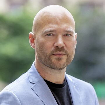 Porträtfoto von Prof. Richard Traunmüller. Er trägt ein hellblaues Sakko, ein schwarzes Shirt, er hat einen Bart. 