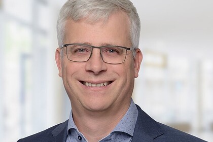 Portrait von Eckhard Janeba. Er trägt ein blaues Hemd sowie einen dunkelblauen Blazer und steht lächelönd vor einer Fensterfront.