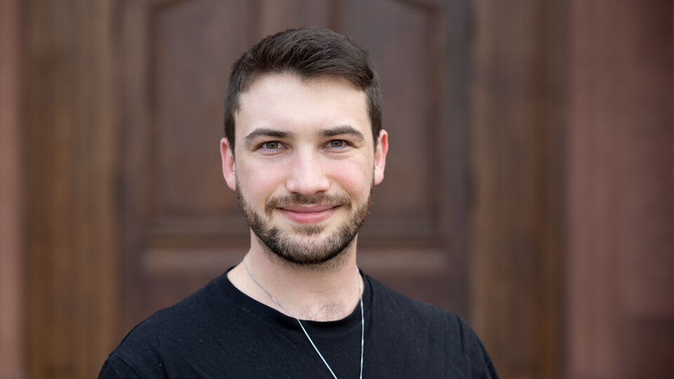 Noam Himmelrath. Er trägt ein schwarzes T-Shirt und steht vor einer dunkelbraunen Holztür.