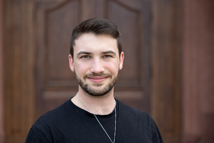Noam Himmelrath. Er trägt ein schwarzes T-Shirt und steht vor einer dunkelbraunen Holztür.