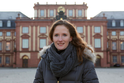Eine Person trägt einen schwarzen Pollover, eine grau Jacke mit Fell sowie einen grauen Schal und steht auf dem Ehrenhof. Die Person heißt Karin Hoisl.